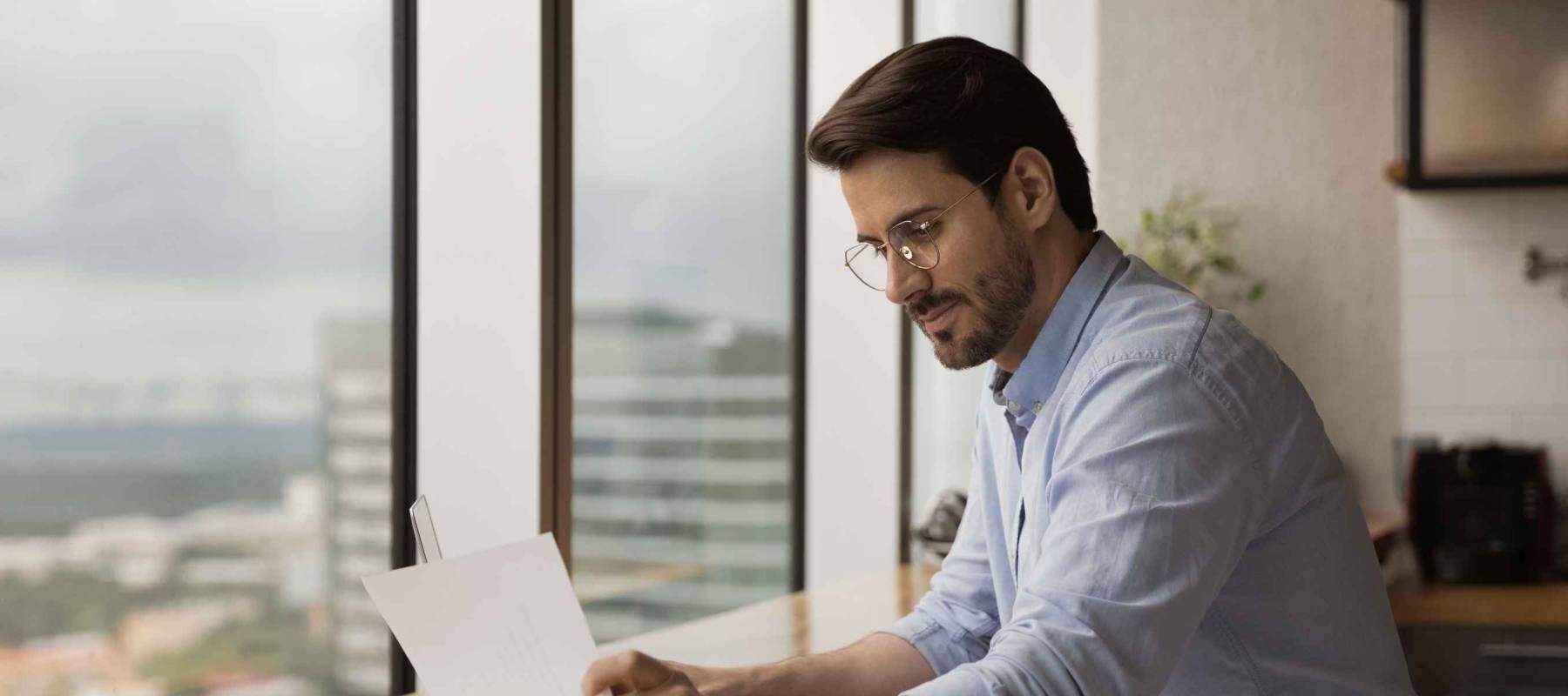 man in blue button up shirt rolled up sleeves holds up paper. He's sitting in front of several windows in a high rise building.