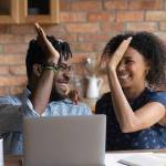 Happy excited Black mixed race couple celebrating financial success at laptop,