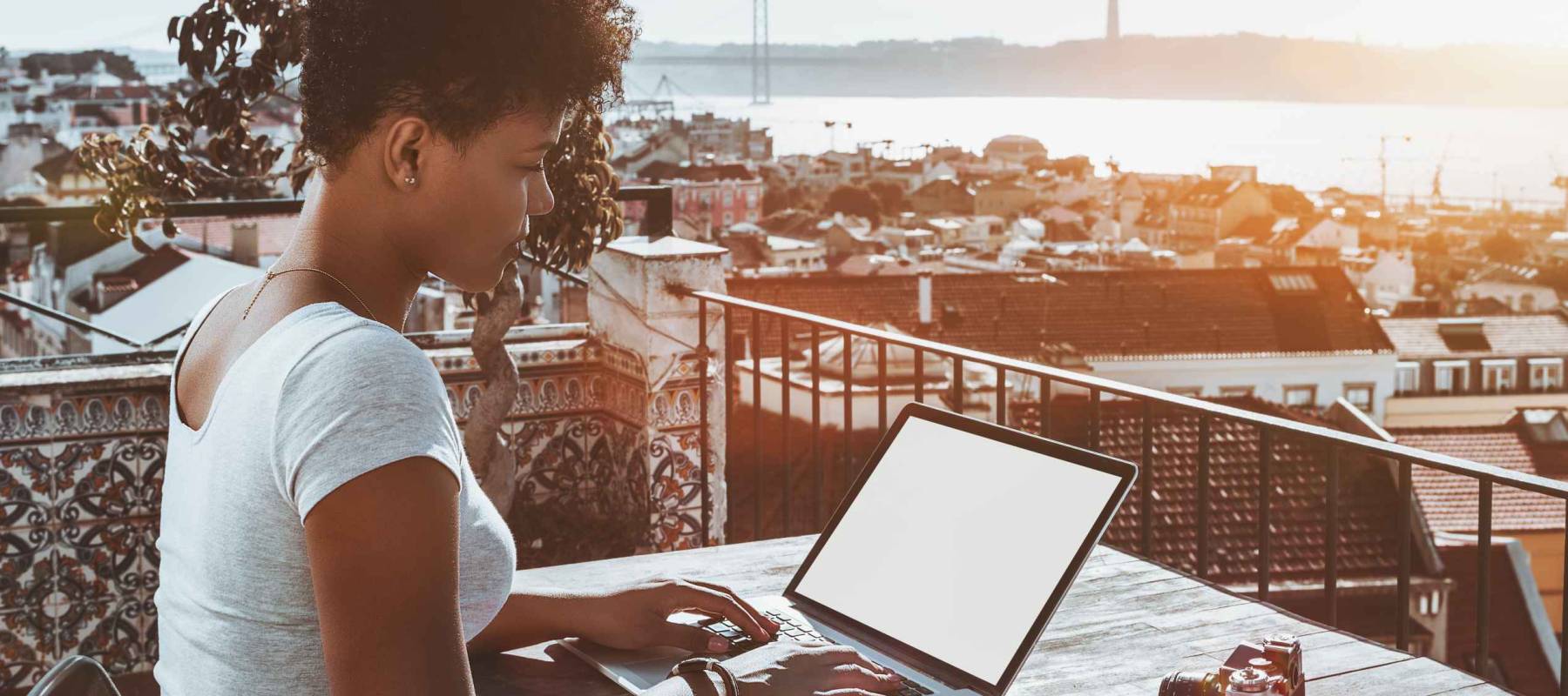 Worker sitting on balcony of Lisbon hotel with stunning bright cityscape in background and working with laptop