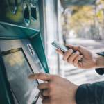 Closeup of male hands using smart phone while typing on ATM, bank machine