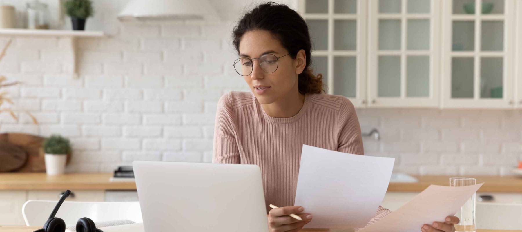 Thoughtful hispanic female sit at home workplace hold financial documents look on pc screen check data.