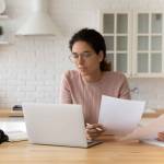 Thoughtful hispanic female sit at home workplace hold financial documents look on pc screen check data.