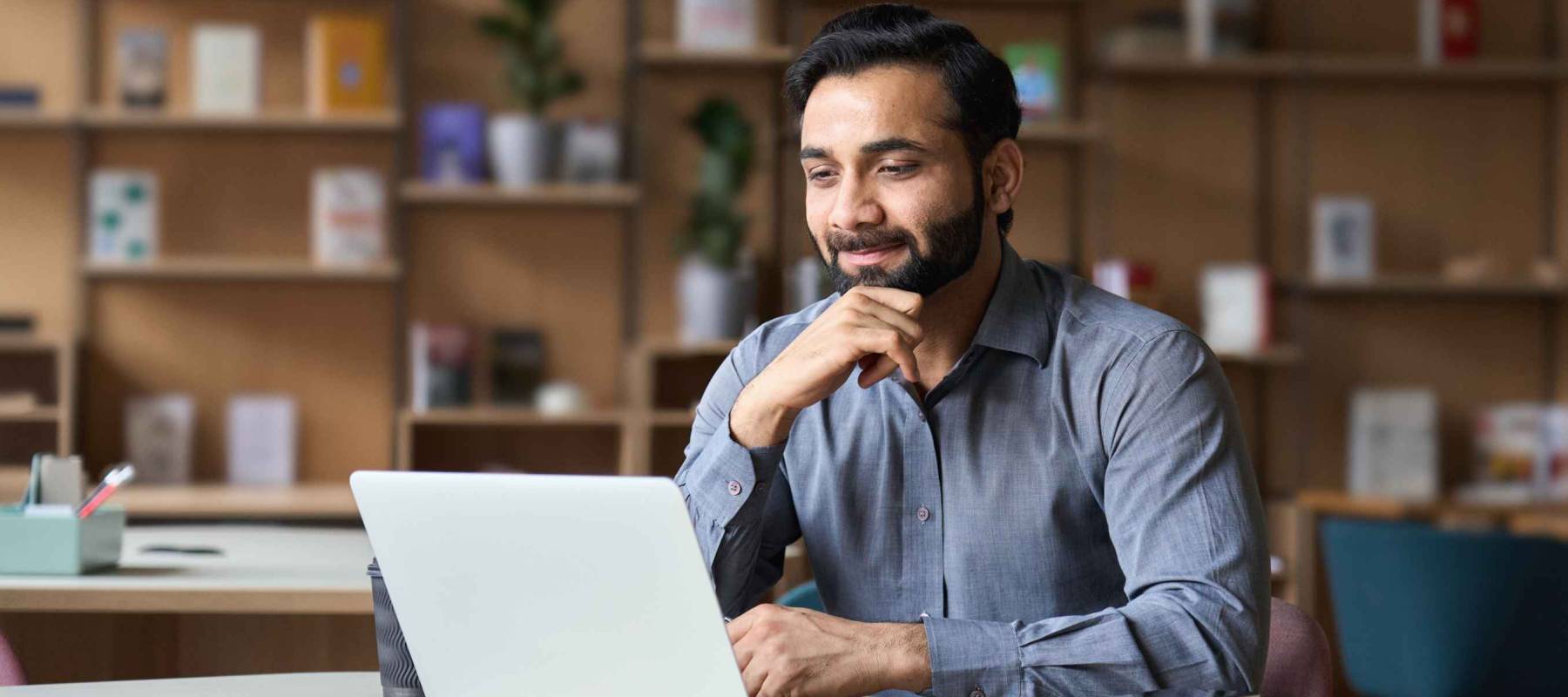Smiling indian businessman working on laptop in modern office lobby space.