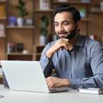 Smiling indian businessman working on laptop in modern office lobby space.