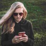 blonde woman standing in a field wearing glasses holding a cellphone wondering why