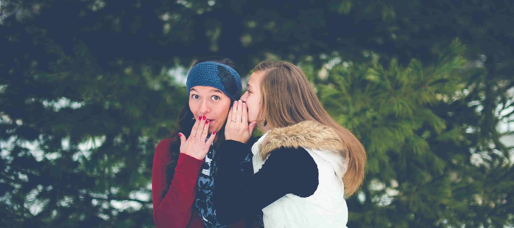 two women in winter in forest. One whispering investment tips in the other's ear. The other is excited by the news.