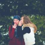 two women in winter in forest. One whispering investment tips in the other's ear. The other is excited by the news.