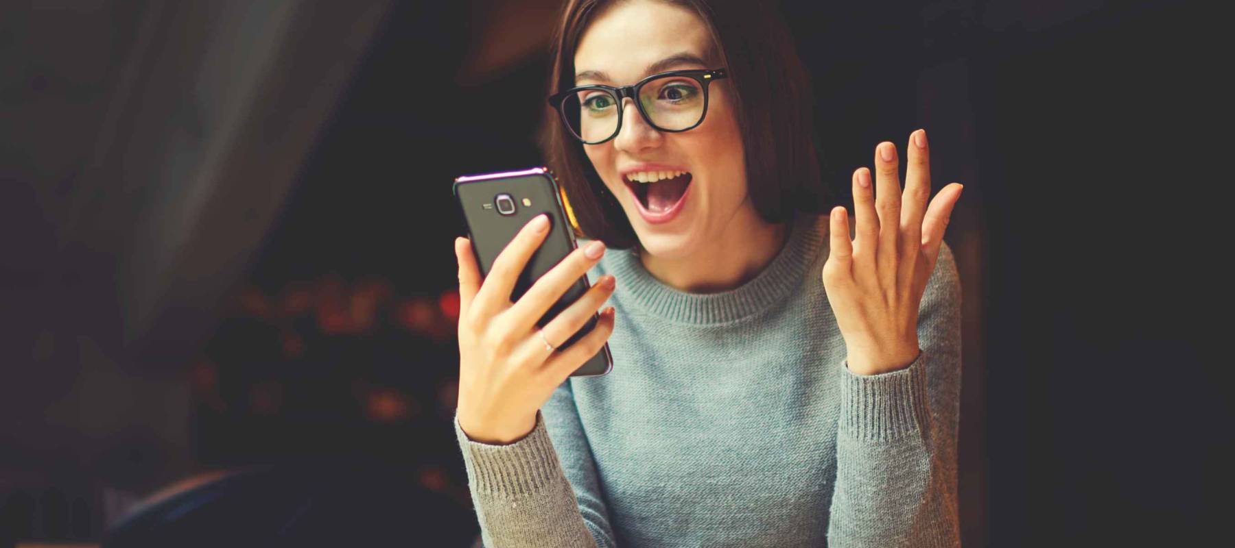 Surprised young female blogger chatting with best friends in social network on mobile phone while received shocking good news