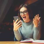 Surprised young female blogger chatting with best friends in social network on mobile phone while received shocking good news