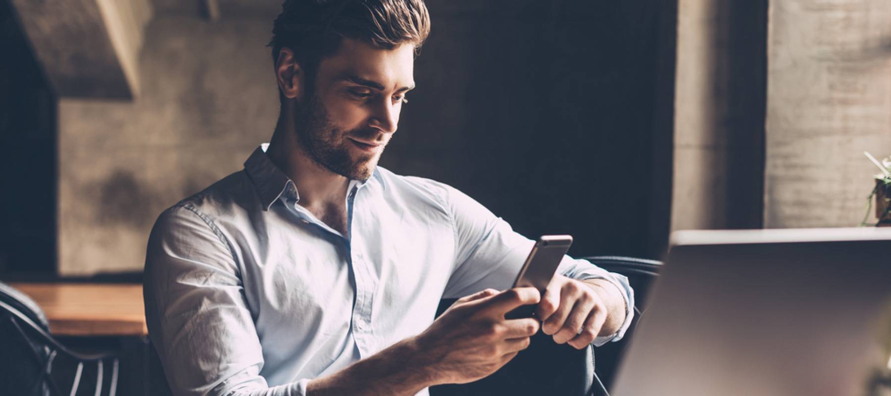 Confident young man in smart casual wear holding smart phone and looking at it while sitting at his working place in office