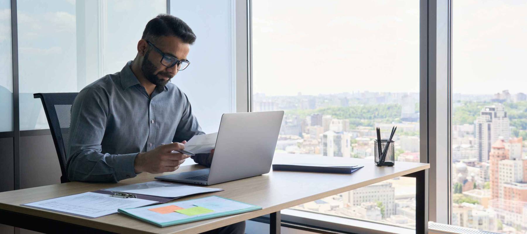 Indian latin focused financier sitting at desk doing paperwork working with bank taxes, loans, debit, credit operations using laptop near panoramic window in contemporary corporation office.