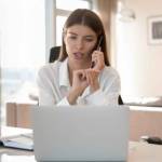 Serious beautiful young freelance professional girl talking to customer on cellphone at home office workplace, giving telephone support, looking at laptop, speaking at table with pc