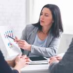 Professional woman broker giving presentation to young couple in her office, showing graphs to explain information to them