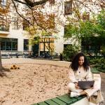 Smiling young african girl wearing autumn coat using laptop while sitting in lotus pose near sandbox