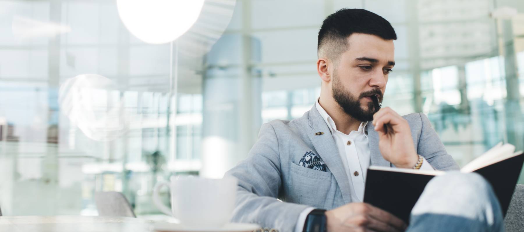 Serious bearded businessman pensively looking down while thinking on work issues during coffee break in modern office with glass walls