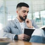 Serious bearded businessman pensively looking down while thinking on work issues during coffee break in modern office with glass walls