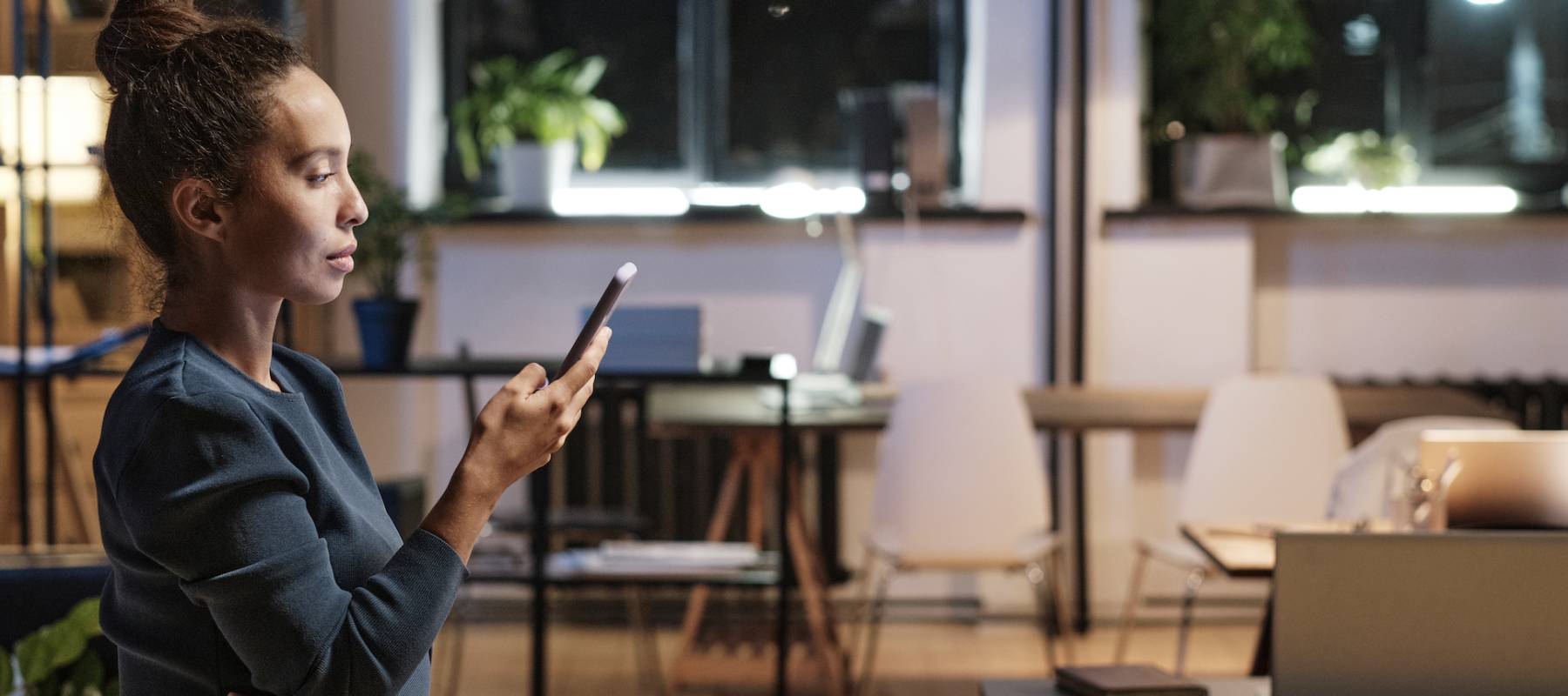 Serious attractive young mixed race businesswoman standing with smartphone in empty office at night