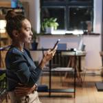 Serious attractive young mixed race businesswoman standing with smartphone in empty office at night
