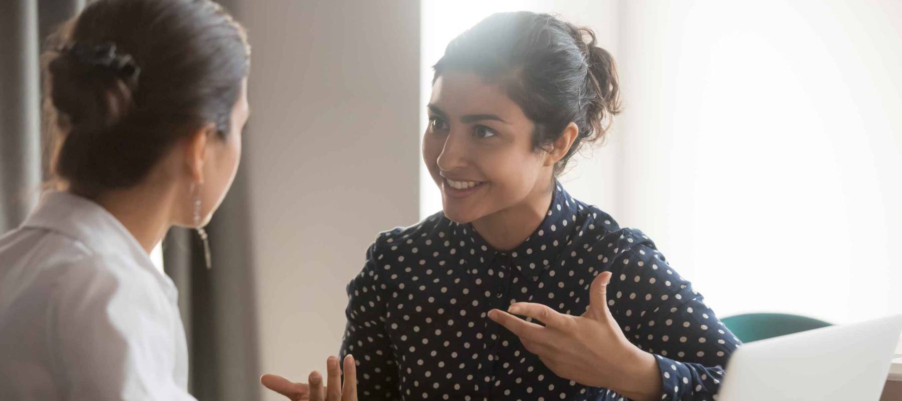 Excited multiethnic female employees discuss work issues sitting at office table, smiling diverse women workers