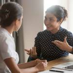 Excited multiethnic female employees discuss work issues sitting at office table, smiling diverse women workers