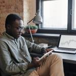 Horizontal medium shot of young Black man sitting at desk in office working with stock trading charts and graphs