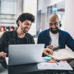 Happy Middle Eastern graphic designers discussing creative sketches during collaborative brainstorming in modern coworking space, cheerful small owners planning together business projecting