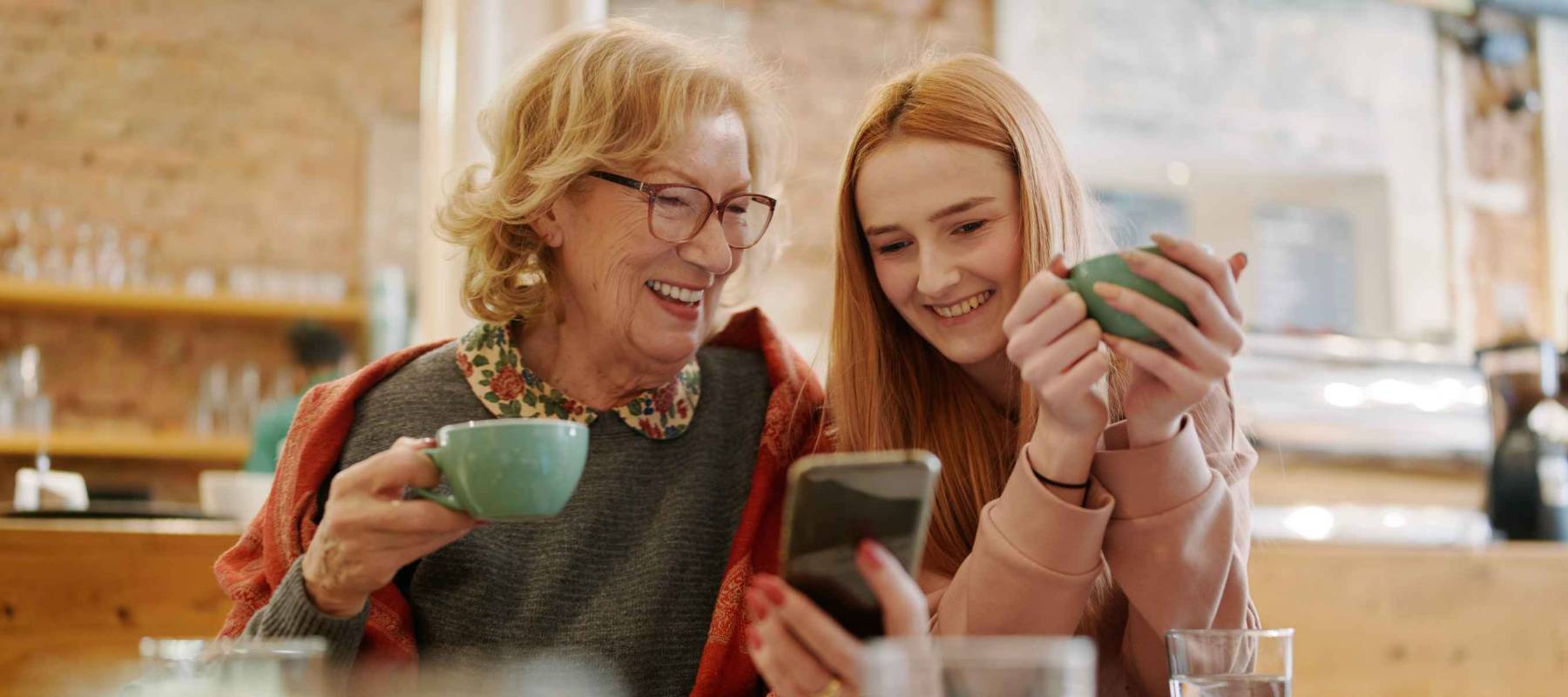 Happy grandmother and her adolescent granddaughter sitting in a cafe, enjoying coffee and using smart phone for funny videos.