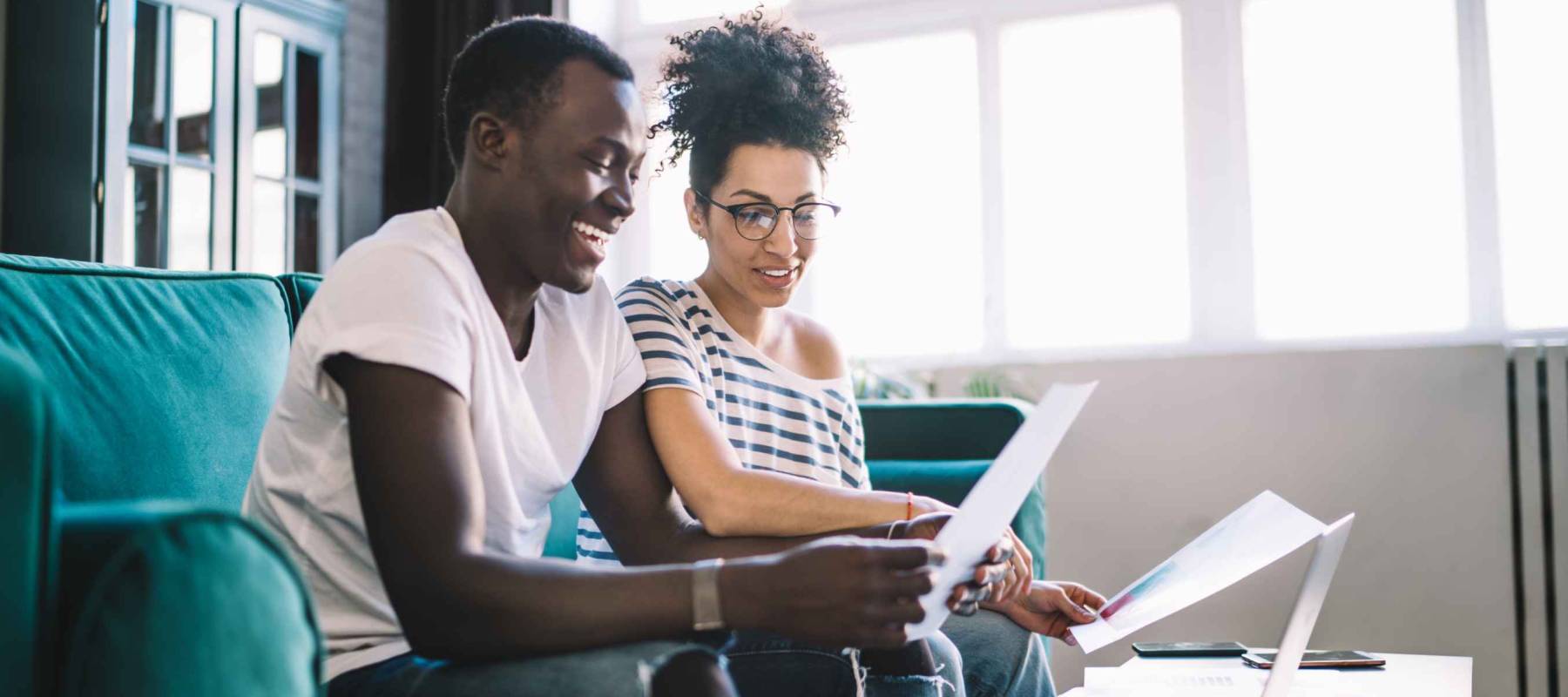 Side view of happy casual multiracial couple relaxing on sofa watching laptop reading paper documents talking and smiling on daytime