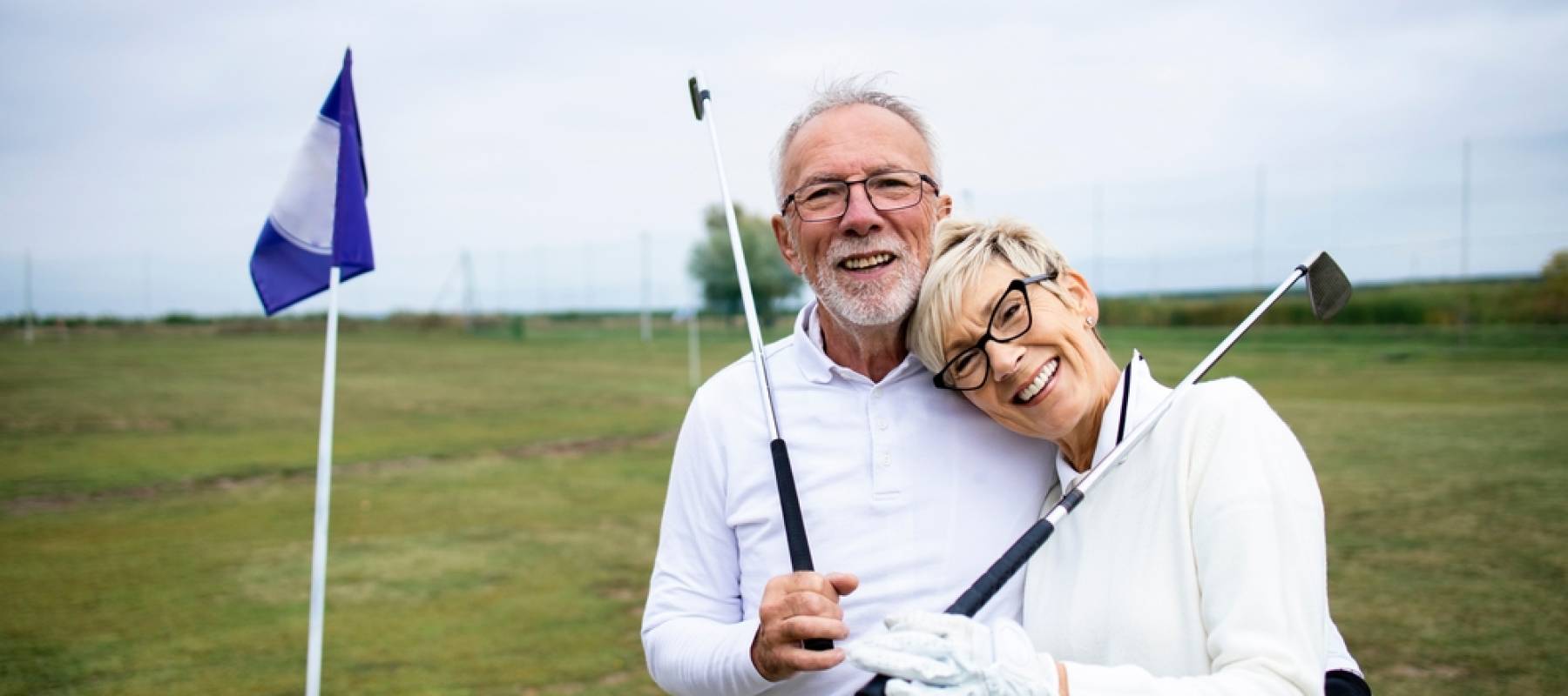 Older couple happily pose holding golf clubs on golf course, smiling at camera.