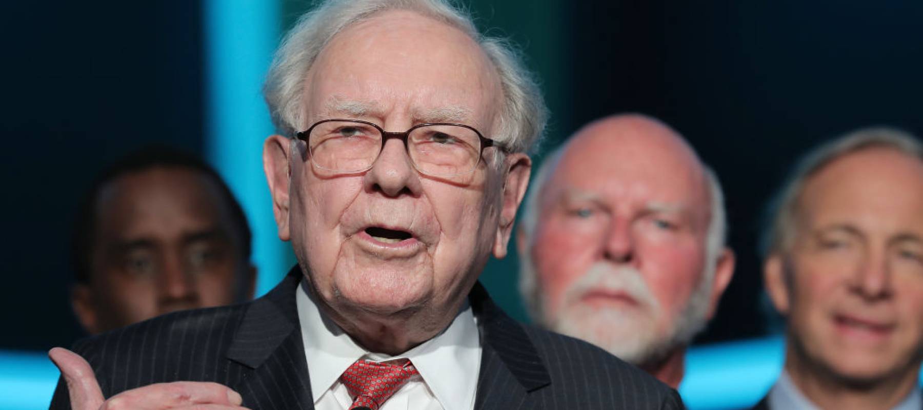 Warren Buffett speaks on stage, making hand gestures.
