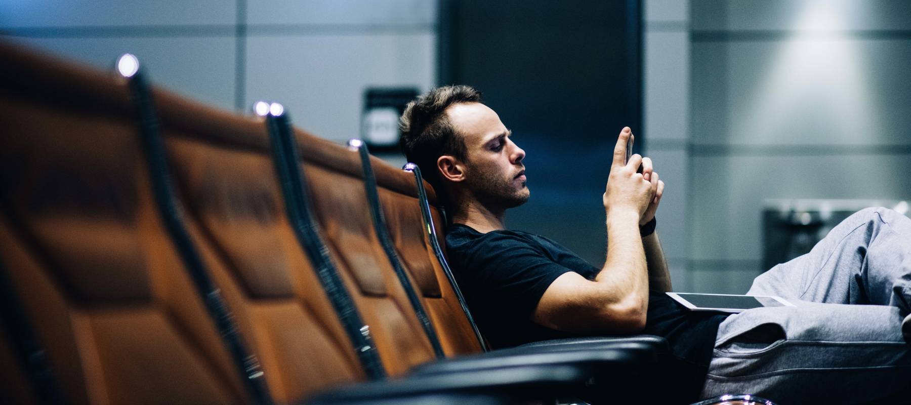 Young man sitting in waiting hall of airport and browsing on his smart phone