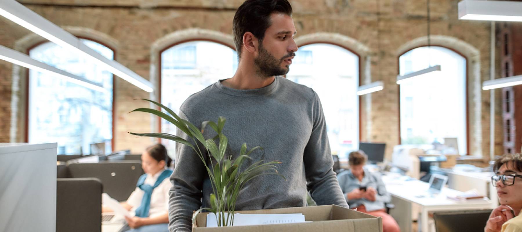 Young man holding box of belongings, looking sad and to the side as he leaves office.