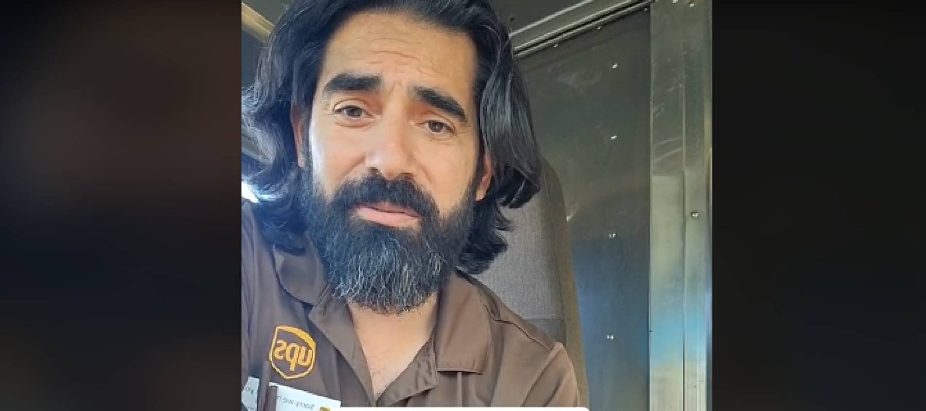 Man in UPS uniform crouches in back of truck talking to camera.