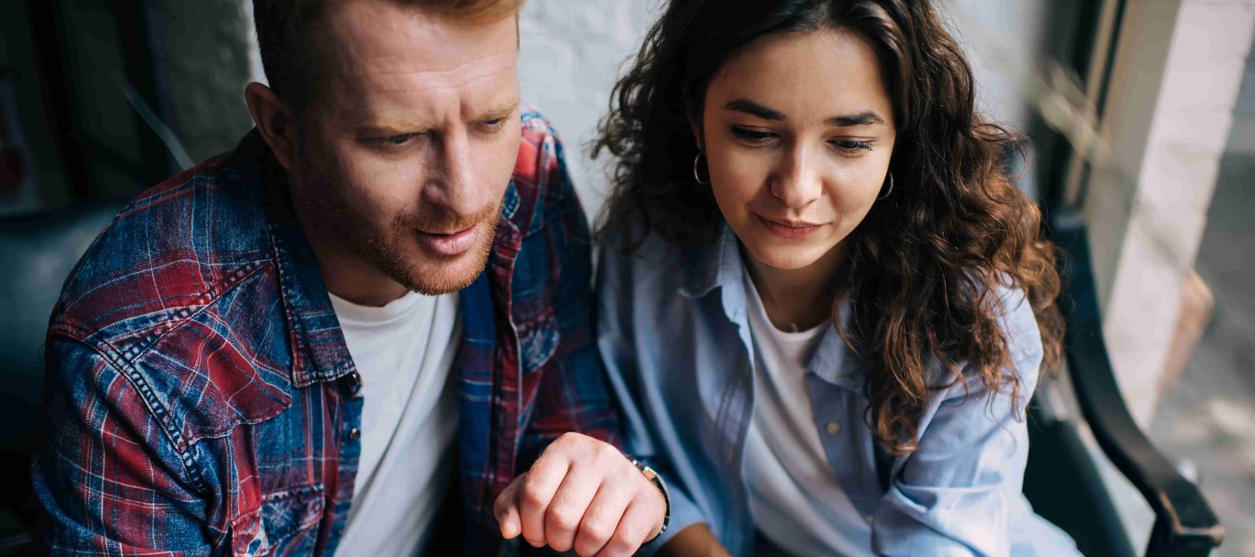 From above of concentrated male freelancer sitting at table with laptop and looking at screen with female friend and reading information showing fingers while working on project in cafe