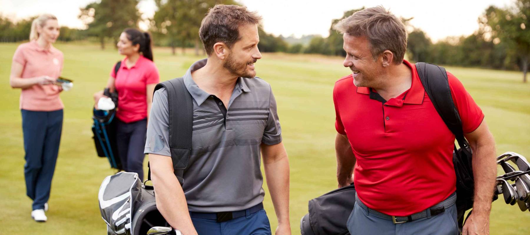 Two Mature Couples Playing Round Of Golf Carrying Golf Bags Along Fairway