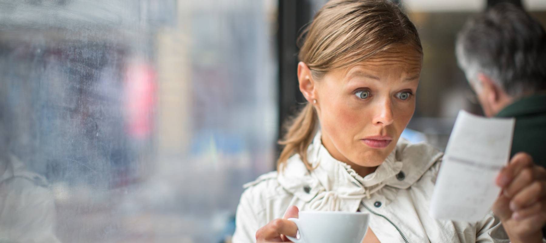 Woman holding cup of coffee looks at bill with surprise.