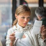 Woman holding cup of coffee looks at bill with surprise.
