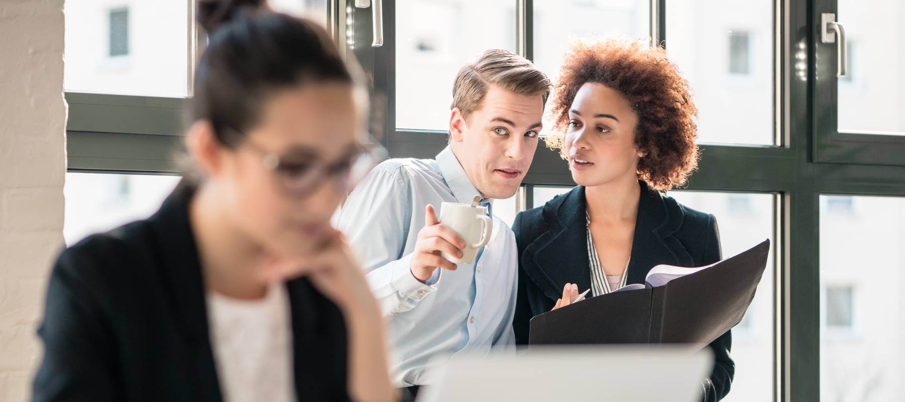 Office employees gossiping in background while woman works on laptop.