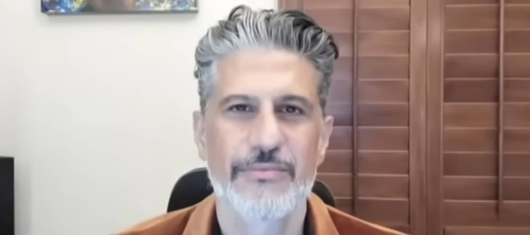 Grey-haired man looking directly at camera, on video chat from home office.