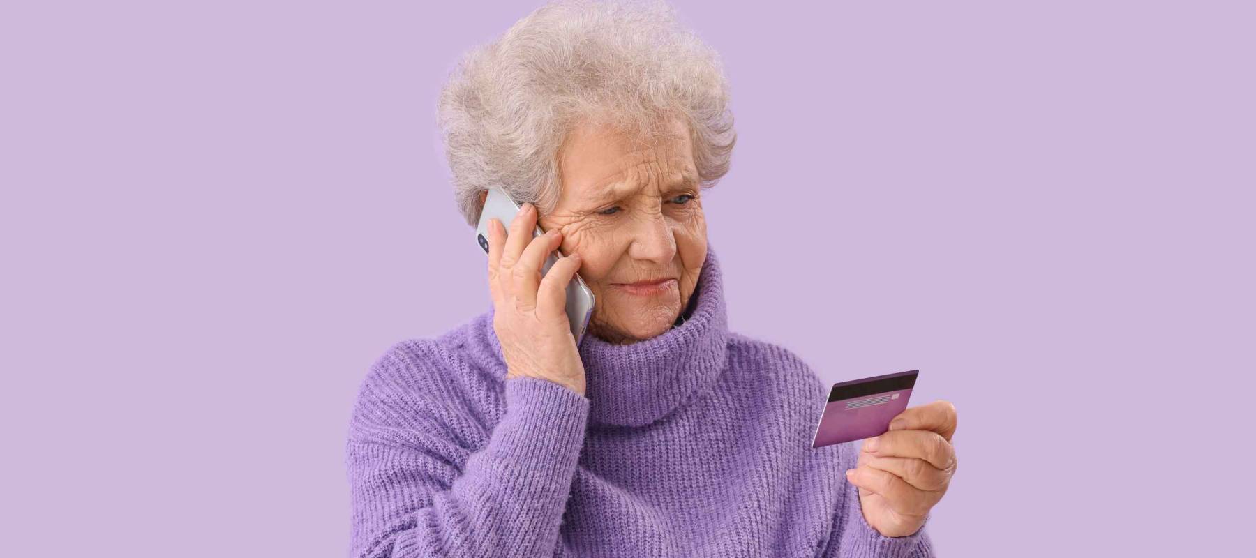 Senior woman with credit card talking by mobile phone on lilac background