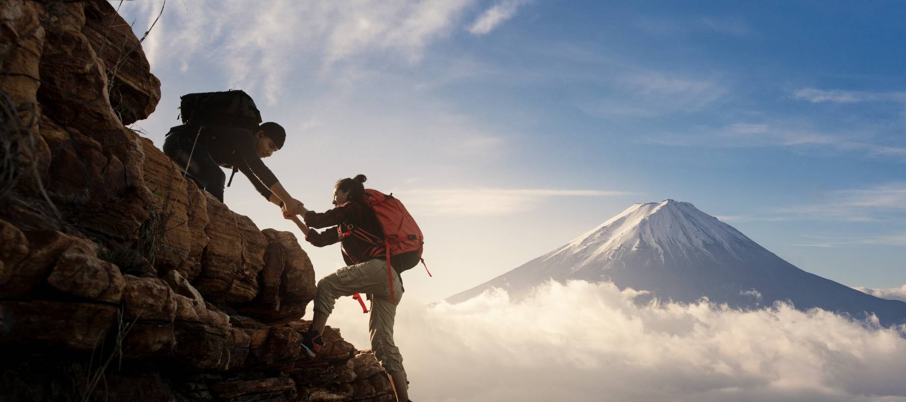 Two mountain climbers, one pulling the other up with a mountain seen in the misty background.