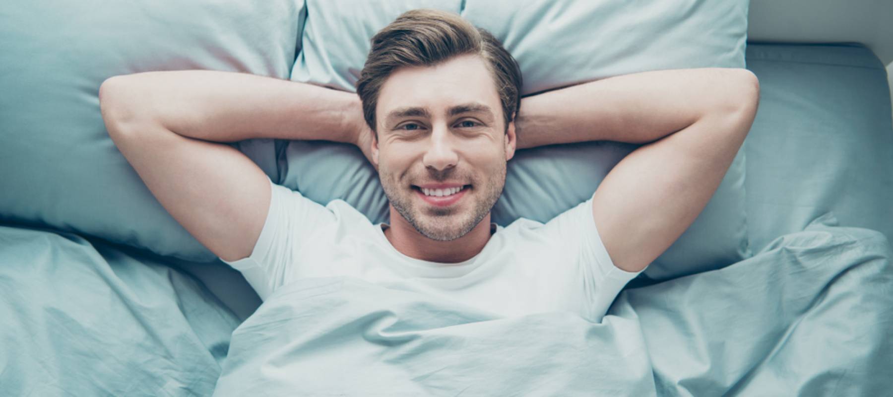 Man lying bed smiling wearing white t-shirt.