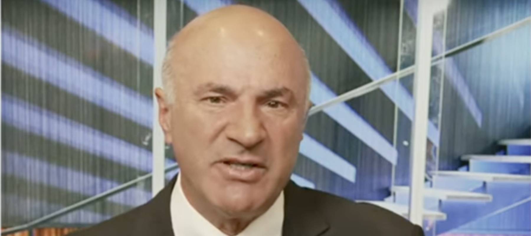Kevin O'Leary on a tv show set, speaking directly to camera with a serious face.