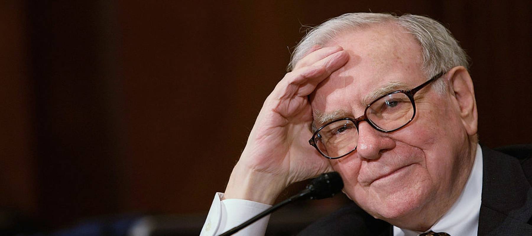 Warren Buffett with a bemused look, sitting in front of a microphone, looking at the camera directly.