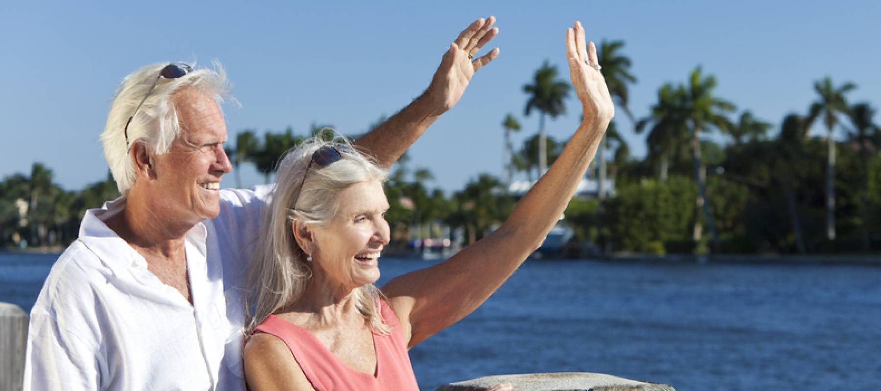 Happy senior couple together outside in sunshine waving by the sea.