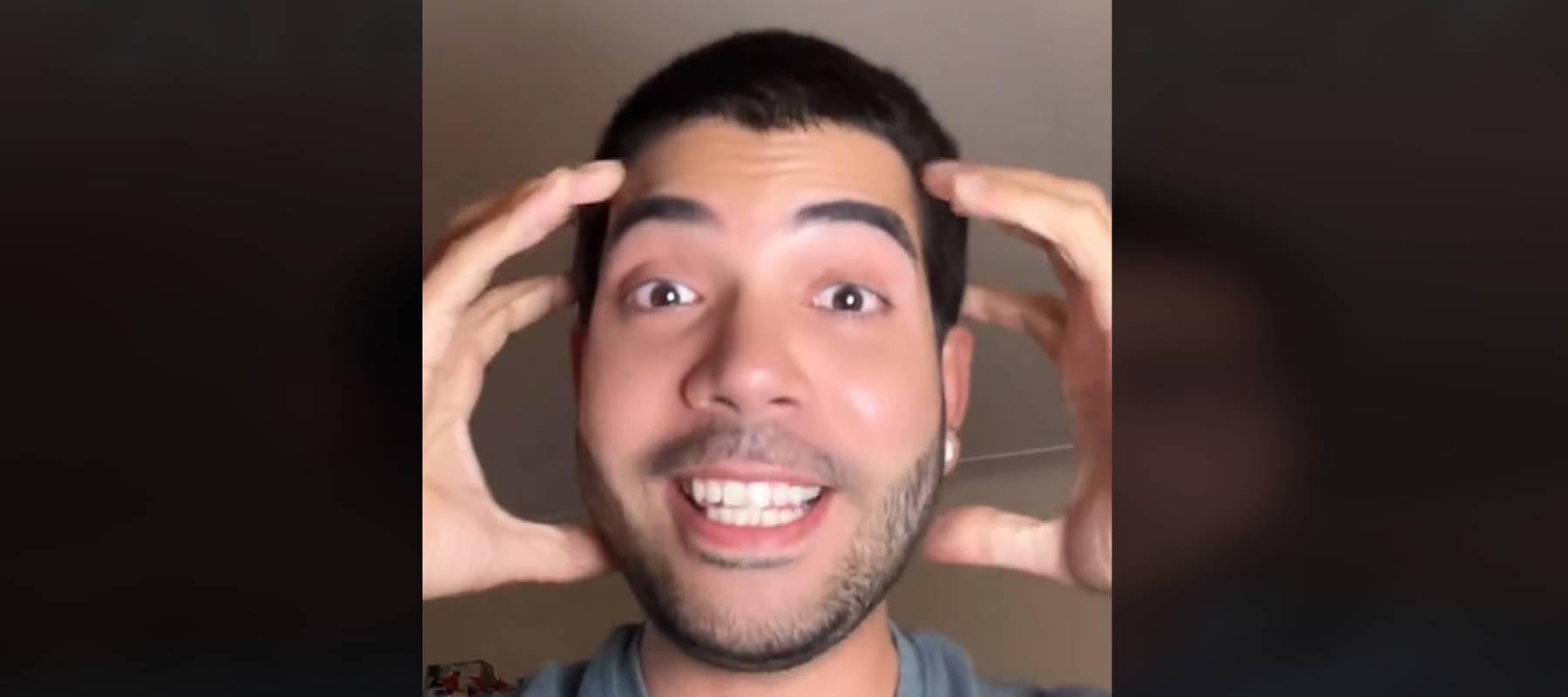 Man seen in his room talking directly to camera, hands held up to his head and making an expression of surprise and delight.
