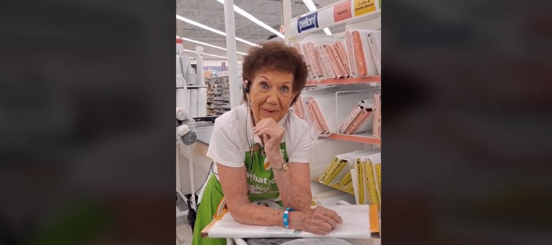 Older woman seen wearing Joann&#039;s Fabrics apron and leaning on table in aisle of the store.