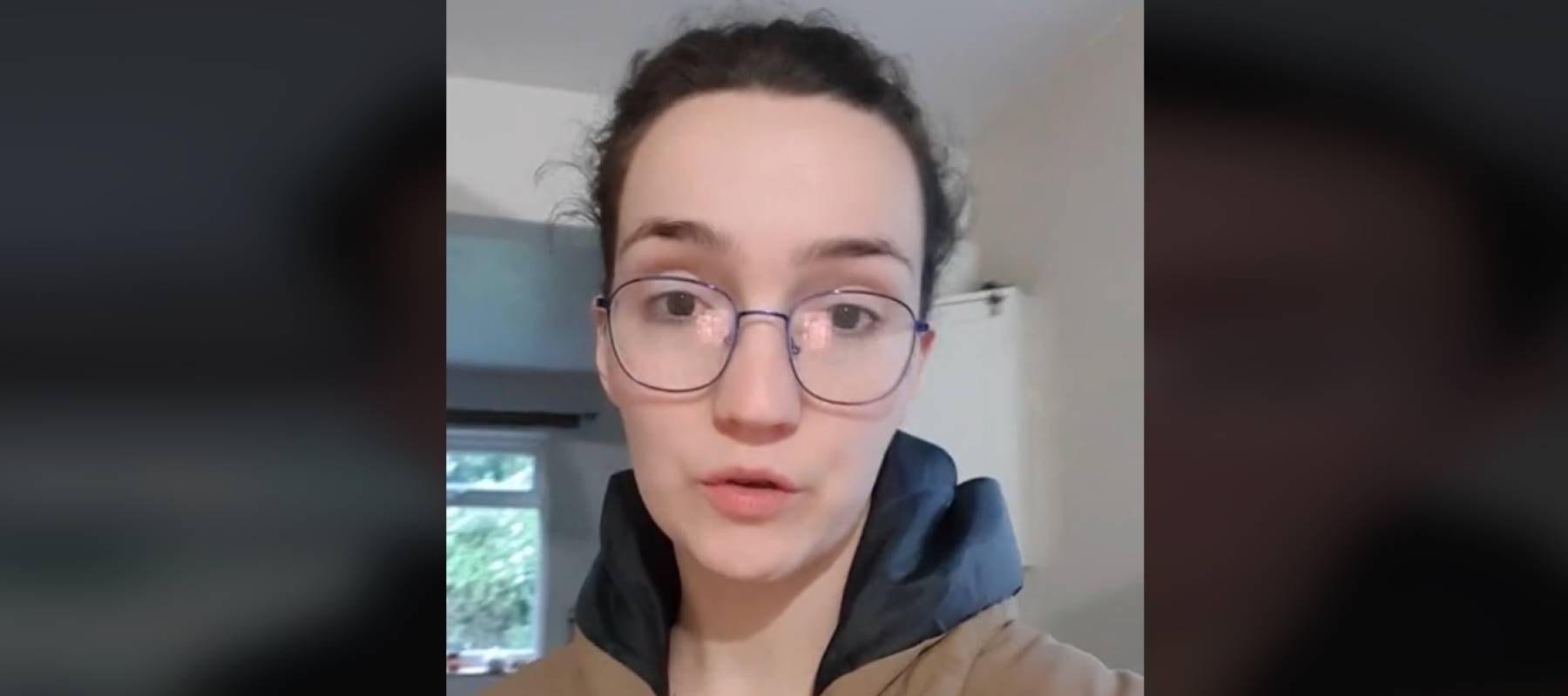 Woman in glasses, wearing coat and swear looking at camera directly while standing in a kitchen.