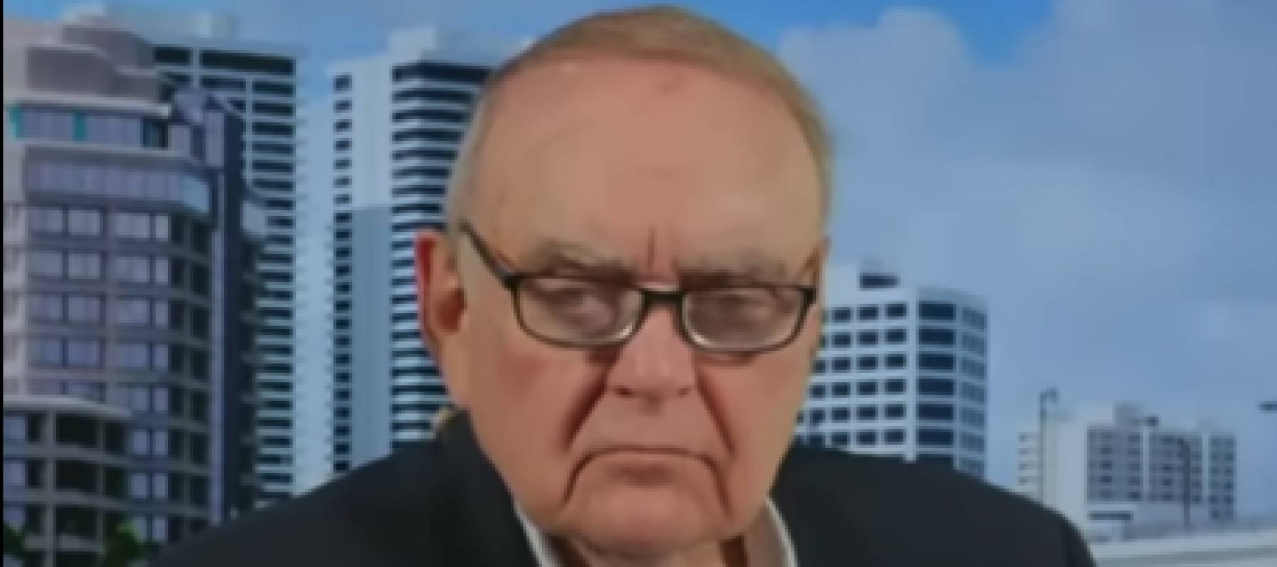 Older man in glasses sits on television set, frowning at the camera.