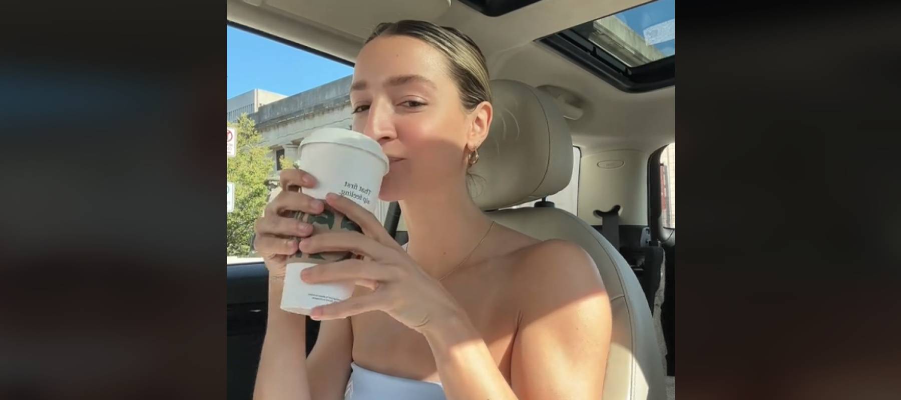 Young woman sits in driver&#039;s side of the car sipping a Starbucks drink and looking at the camera.
